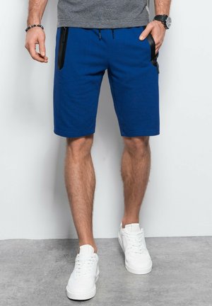 Shorts bleus en tissu doux, avec des poches latérales noires zippées et une taille ajustable à cordon. Portés avec des baskets blanches.