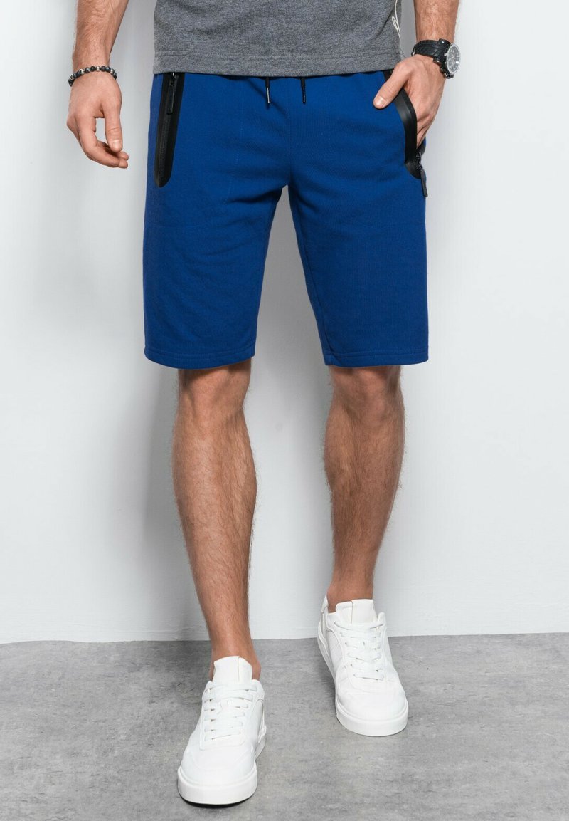 Pantaloni corto blu realizzati in un tessuto morbido, con tasche laterali nere con cerniere e vita regolabile con cordino. Indossati con sneakers bianche.