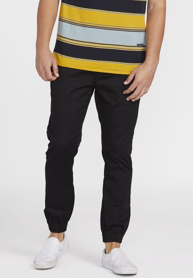 Homme portant un pantalon de jogging noir, des chaussures blanches sans lacets et une chemise rayée jaune, noire et bleu clair sur un fond uni.