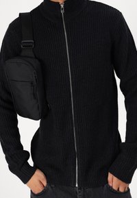 Svart ribbad stickad tröja med hög krage och full dragkedja, kombinerad med en svart axelremsväska med en dragkedjeförsedd ficka.