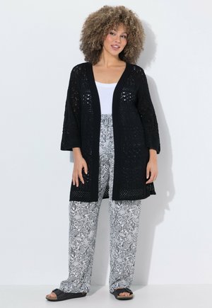 Donna con capelli ricci indossa un cardigan nero a maglia aperta, top bianco, pantaloni ampi con motivo e sandali neri aperti, in piedi contro una parete bianca.
