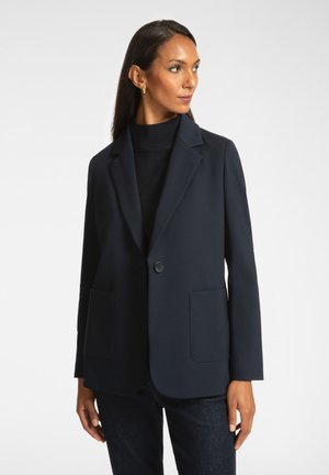 Marineblazer van gladde stof, met een notch lapel, enkele knoopsluiting en twee voorzakken. Gecombineerd met een zwarte coltrui.