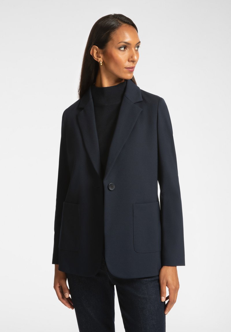 Granatowa marynarka uszyta z gładkiego materiału, z wyciętym klapem, zapinana na jeden guzik, z dwiema przednimi kieszeniami. Połączona z czarnym golfem.