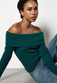Giovane donna dai capelli ricci seduta contro un muro bianco, che indossa un maglione azzurro off-shoulder e jeans blu, mentre guarda tranquillamente di fronte a sé.