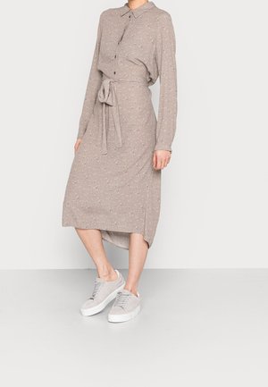 Kobieta w beżowej sukience midi z długim rękawem, z małym kwiatowym wzorem, z wiązaną kokardą w talii i jasnoszarych sneakersach na platformie.
