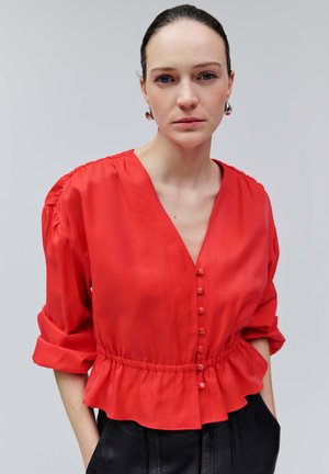 Femme aux cheveux foncés relevés, portant des boucles d'oreilles argentées en anneau, un chemisier rouge boutonné avec une taille à volants, et un pantalon noir, les mains dans les poches.