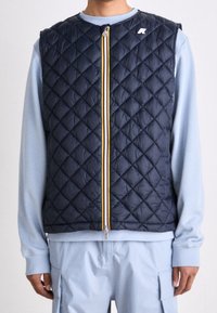 Marineblaue gesteppte Weste mit einem vorderen Reißverschluss, der gelbe und weiße Streifen aufweist. Kombiniert mit einem hellblauen Langarmshirt und passenden Hosen.