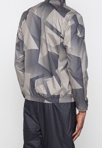 Lekka, wzorzysta kurtka w szarym kolorze z ciemnymi geometrycznymi wzorami. Posiada elastyczne mankiety i dół, wykonana z gładkiego syntetycznego materiału.