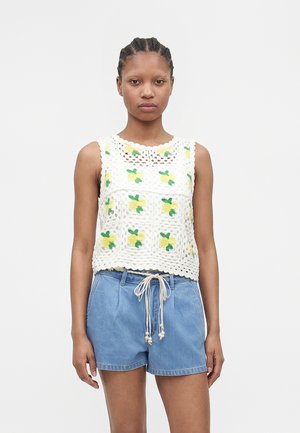 Giovane donna con i capelli intrecciati indossa un top bianco all'uncinetto con fiori gialli e pantaloncini di jeans blu con vita regolabile.