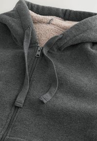Felpa grigia con zip caratterizzata da un esterno testurizzato, rivestimento interno rosa chiaro, doppio cordino e zip frontale.