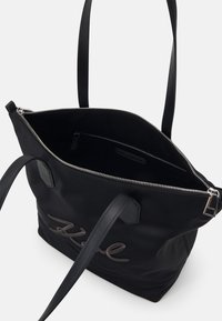 Svart tote-bag med två remmar, öppen silverdragkedja, synligt dragkedjefack inuti och broderad kursiv text på framsidan.