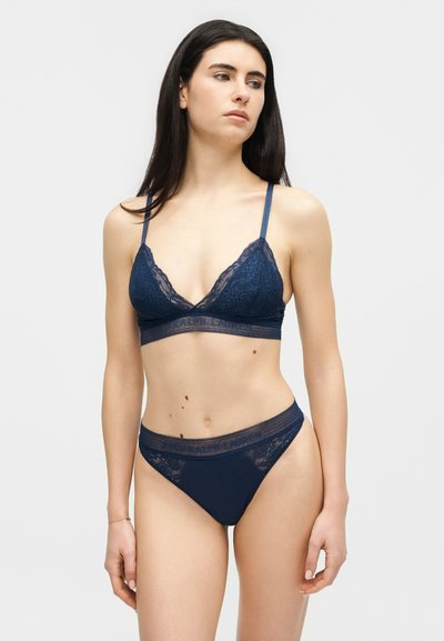 Γυναίκα ντυμένη με μπλε κοντό σουτιέν από δαντέλα και ταιριαστά ψηλοκάβαλα εσώρουχα, στέκεται μπροστά σε απλό φωτεινό φόντο, κοιτάζοντας ελαφρώς προς το πλάι.