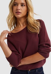 Bordeaux geribbelde trui met een off-the-shoulder ontwerp en opgerolde mouwen. Zachte textuur met zichtbare horizontale strepen. Casual pasvorm.