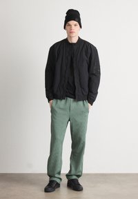 Schwarze Bomberjacke aus strukturiertem Stoff, kombiniert mit grünen gemusterten Jogginghosen. Schwarze Sneaker runden das Outfit ab, ergänzt durch eine schwarze Mütze.