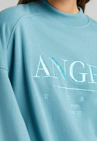 Ljusblå sweatshirt med ribbad halsringning och broderad text. Har en avslappnad passform och mjuk textur, med ytterligare textdetaljer.