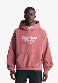 Pink hooded sweatshirt with front pocket and white text "Cold Heart Theory." Features relaxed fit and soft texture. Worn with dark jeans.