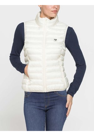 Donna che indossa un gilet bianco trapuntato sopra una maglia a maniche lunghe blu navy e jeans blu, in piedi con una mano nella tasca del gilet.