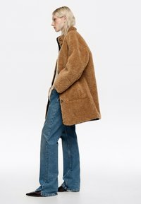 Mujer de pie de lado, vistiendo un abrigo de peluche color beige, jeans de pierna ancha azules y zapatos negros de punta, frente a un fondo blanco.