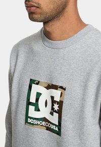 Sudadera gris con un logo de DC en blanco con patrón de camuflaje, con cuello de canalé y tejido texturizado. Diseño ajustado.