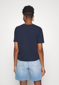 Marinblå bomullströja med korta ärmar, rundad halsringning och avslappnad passform, tillsammans med ljusa denimshorts, vy från baksidan.