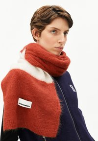 Jeune personne avec les cheveux bruns courts portant une veste zippée bleu marine et une épaisse écharpe en laine orange et blanche étiquetée "ARMEDANGELS."