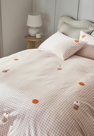 Beige kariertes Bettzeug mit bestickten Kürbissen und Cupcakes auf einem Bett mit gebogenem Kopfteil neben einem hölzernen Nachttisch mit Lampe.