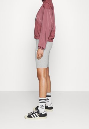 Person standing sideways wearing a mauve jacket, gray bicycle shorts, white Adidas socks, and black Adidas sneakers.