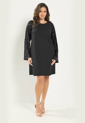 Femme portant une robe noire mi-longue aux manches longues pailletées et des sandales à talons beiges, debout devant un fond uni.
