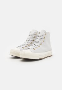 Høye sneakers i lysegrå kanvas med gulløyelletter, hvit gummitopp, teksturert såle, og flere horisontale striper på midtsålen.