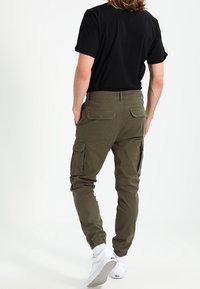 Olive green cargo pants with elastic cuffs, multiple side pockets, and a smooth cotton fabric. Worn with a black shirt and white sneakers.