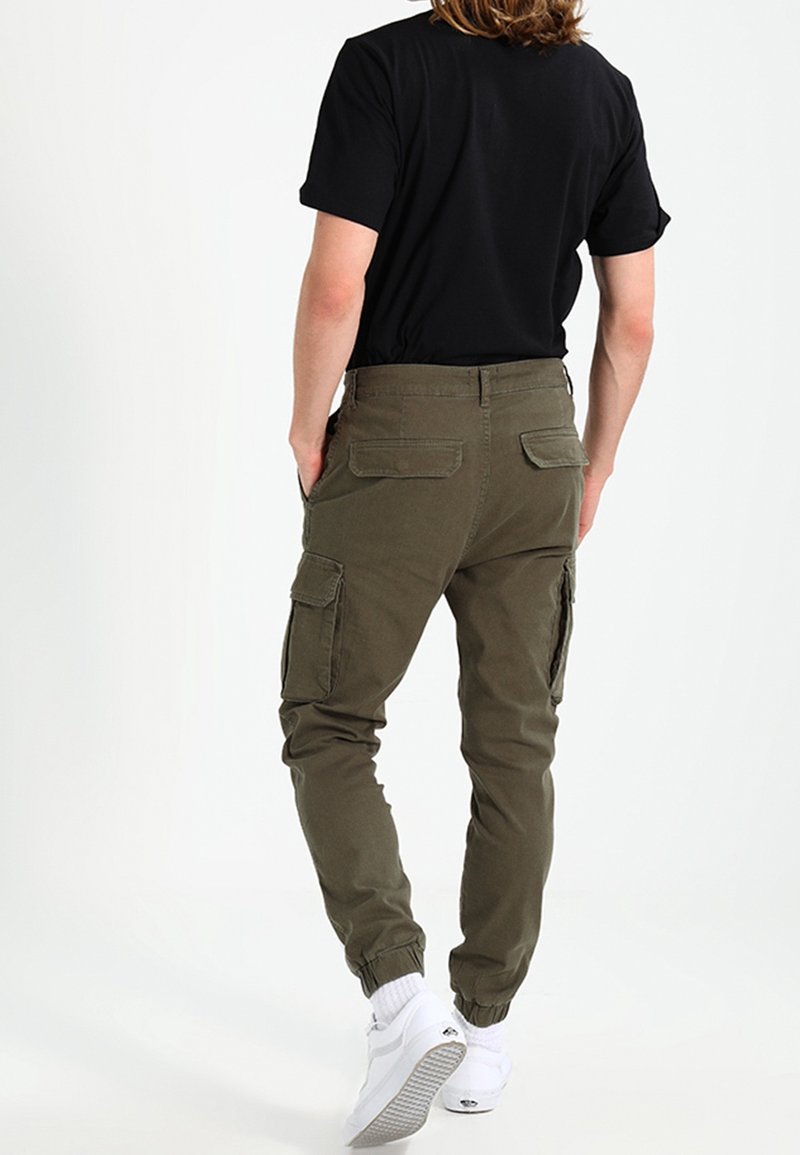 Olive green cargo pants with elastic cuffs, multiple side pockets, and a smooth cotton fabric. Worn with a black shirt and white sneakers.