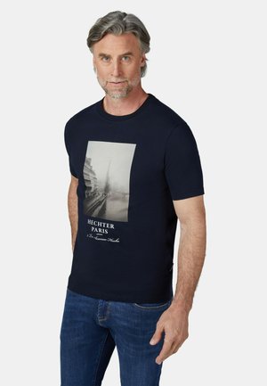 Man met grijs haar en baard draagt een donkerblauw T-shirt met een stadsgezicht van Parijs en tekst, gecombineerd met blauwe spijkerbroek, staand tegen een witte achtergrond.