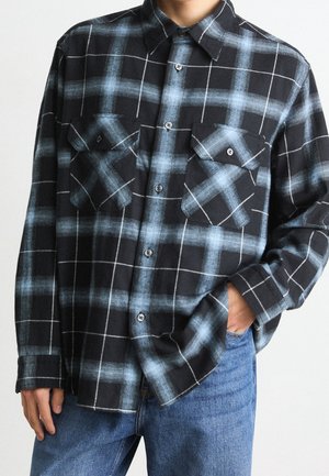 Chemise à carreaux noire et bleue à boutons avec deux poches poitrine portée avec un jean bleu, une main dans la poche, sur une personne recadrée au niveau du cou.