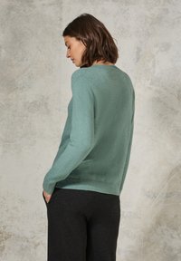 Hellgrüner Pullover aus weichem Material, mit geripptem Saum und Bündchen. Kombiniert mit dunkelgrauen Hosen, die eine lockere Passform und Struktur aufweisen.