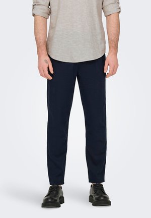 Bărbat poartă o cămașă bej cu mâneci lungi suflecate, pantaloni bleumarin mulați și pantofi negri cu șireturi, stând în picioare pe fundal alb.