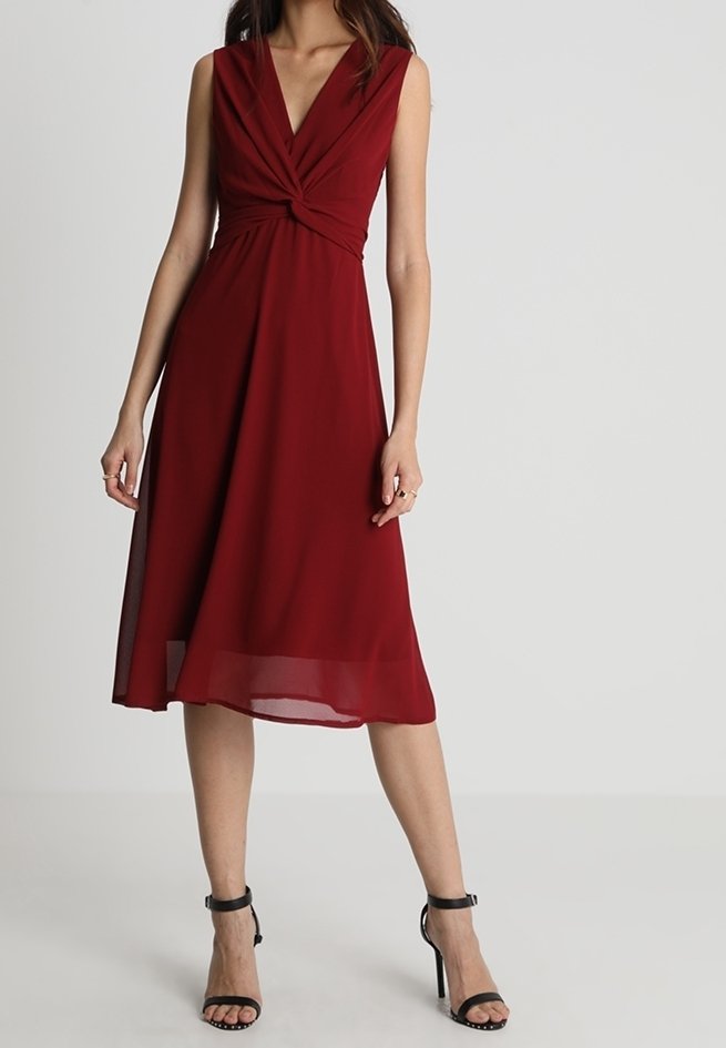 Robe bordeaux sans manches avec un décolleté en v torsadé, tissu fluide, ourlet au niveau du genou, portée avec des escarpins noirs à bride à la cheville.