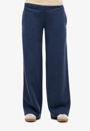 Marineblå vidbundne joggingbukser med elastik i taljen, båret med beige sneakers og en cremefarvet top med blonder synlige ved taljen.