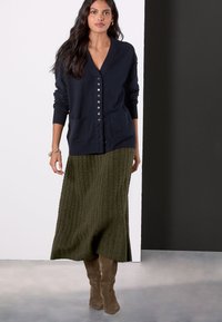 Navy cardigan with V-neck, full-button closure, and two front pockets, paired with an olive green textured knit skirt and brown knee-high boots.