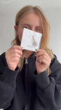 Furla atslēgu piekariņš, kas izstādīts uz baltas kartes; tajā ir sudraba toņa ziedu dizains un zelta sprādze, kas izceļ glancētu apdari.