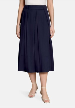 Femme portant une jupe midi bleu marine avec des plis devant, associée à des sandales beiges à talons hauts et à brides sur fond blanc.
