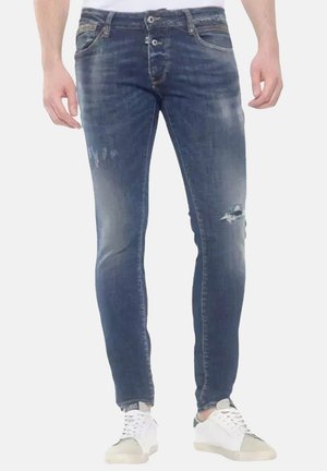 Jeans de mezclilla azul deslavados con un corte ajustado. Presentan detalles desgastados, dos bolsillos frontales y dos traseros. Combinados con zapatillas blancas.