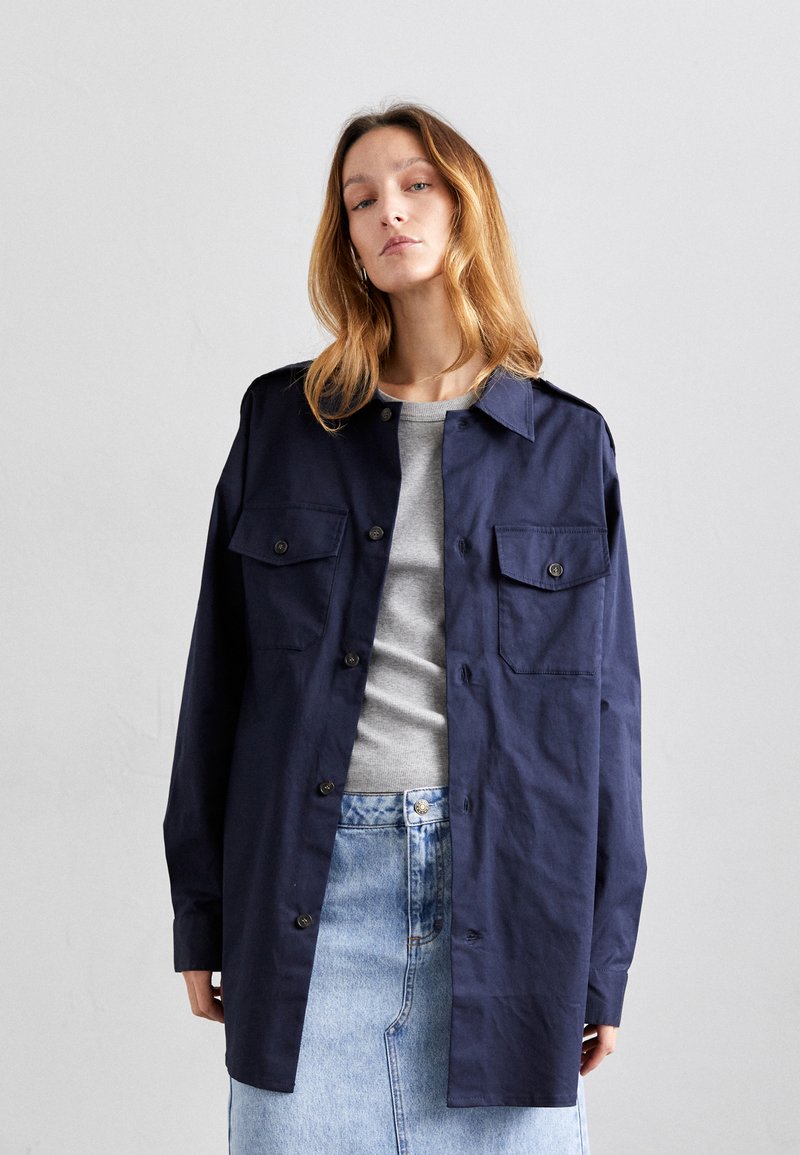 Marineblauwe shirtjack van katoen, met twee voorzakken, knoopsluiting en een ontspannen pasvorm. Draag het over een grijze top en een denimrok.
