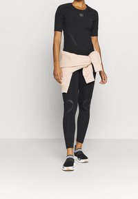 Kobieta w czarnym stroju sportowym z krótkim rękawem, czarnych legginsach, beżowym sweterku związanym w pasie i czarnych trampkach.