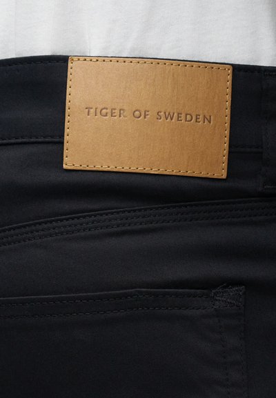 Μαύρο υφασμάτινο παντελόνι με καφέ δερμάτινη ετικέτα αποτυπωμένη με το "TIGER OF SWEDEN." Απαλή υφή με διακριτές λεπτομέρειες ραφής.