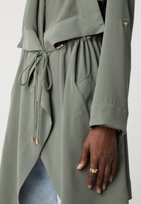 Main à la peau foncée portant une grosse bague en or reposant contre un manteau vert sauge avec taille à cordon et poches avant, sur un jean bleu clair.