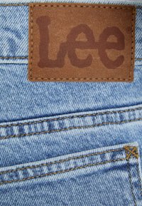 Šviesiai mėlynas džinsinis audinys su rudai odiniu žetonu, ant kurio yra įspaustas "Lee" logotipas. Matomi siuvimo siūlai ir kišenė su pleistru.