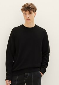 Maglione nero con texture, collo rotondo e maniche lunghe, presenta un orlo a coste. Indossato sopra jeans scuri, design minimalista.