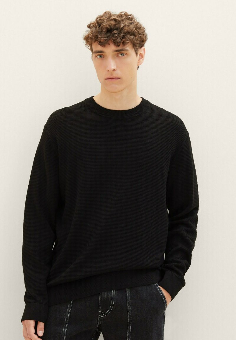 Maglione nero con texture, collo rotondo e maniche lunghe, presenta un orlo a coste. Indossato sopra jeans scuri, design minimalista.