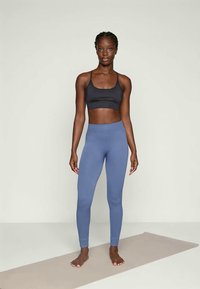 Bustieră sport neagră și colanți albaștri cu dungi pe o siluetă în picioare, desculță, pe un fundal neutru; texturi netede ale materialului vizibile.