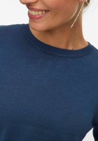 Femme souriante portant un pull en maille bleu et une longue boucle d'oreille en chaîne argentée, visible du cou à la poitrine sur un fond uni.